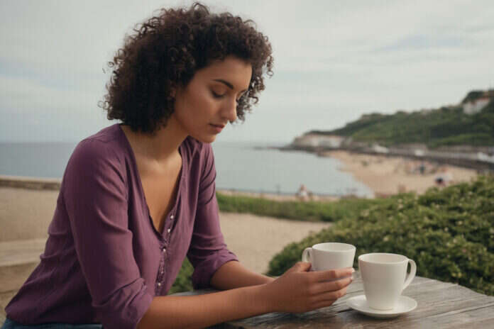 Woman with short curls holding a plain cup beside the coast.